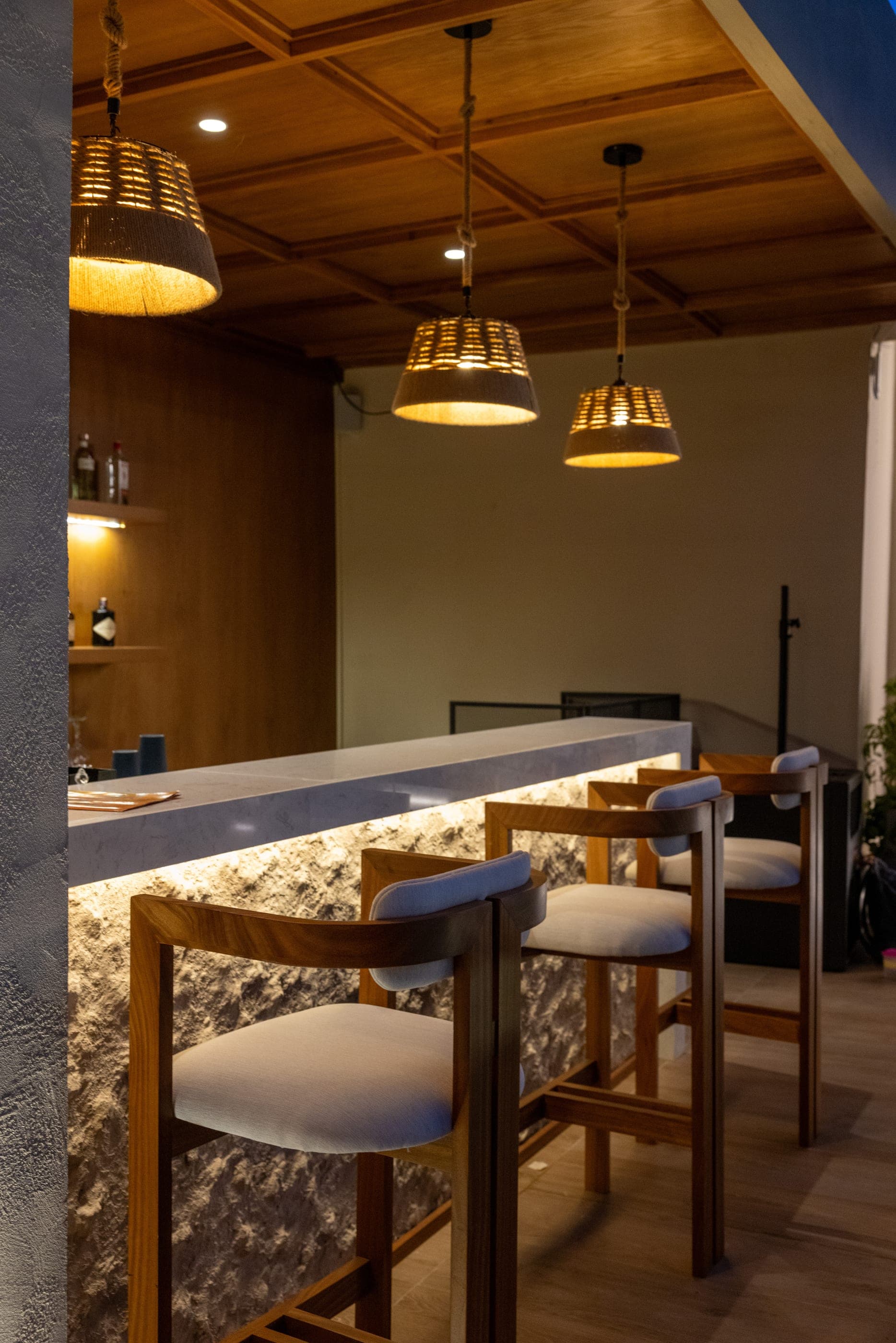 The bar counter in backlit stone, timber stools and woven pendant shades above