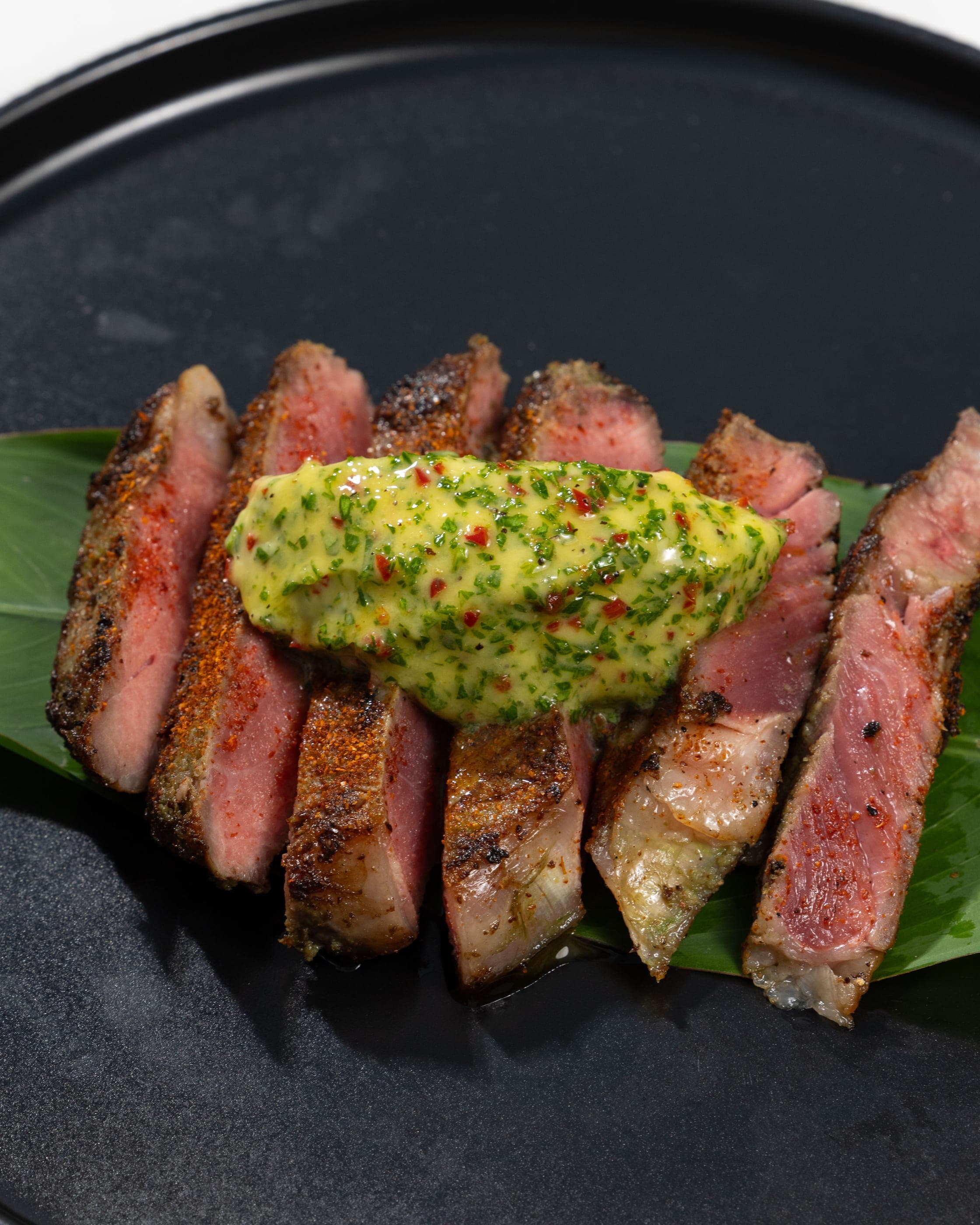Sliced seared steak on a banana leaf under a green herb butter