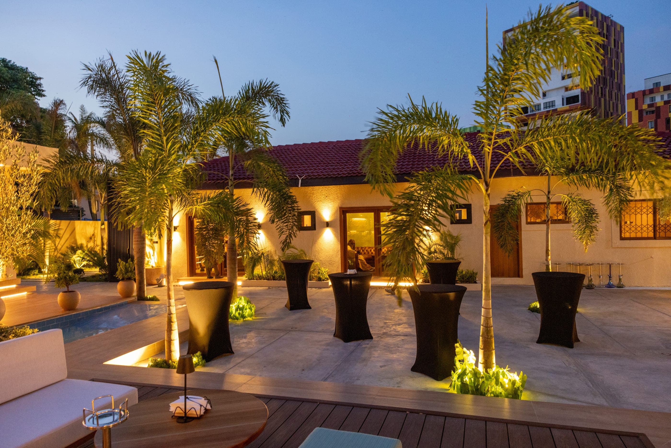 High tables set around the pool at dusk, palms lit from below