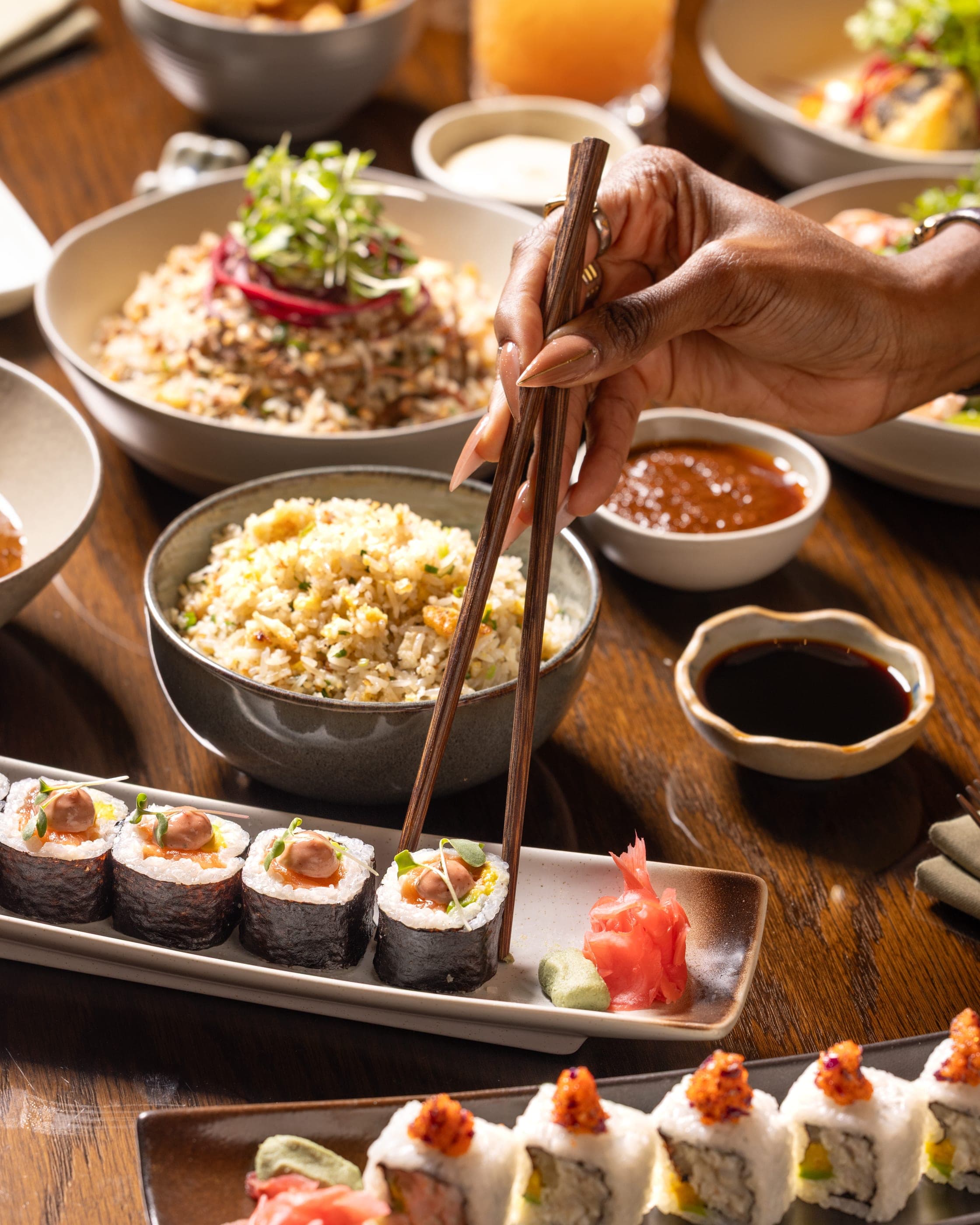 Two pairs of hands reaching across a shared table of rice bowls and sushi
