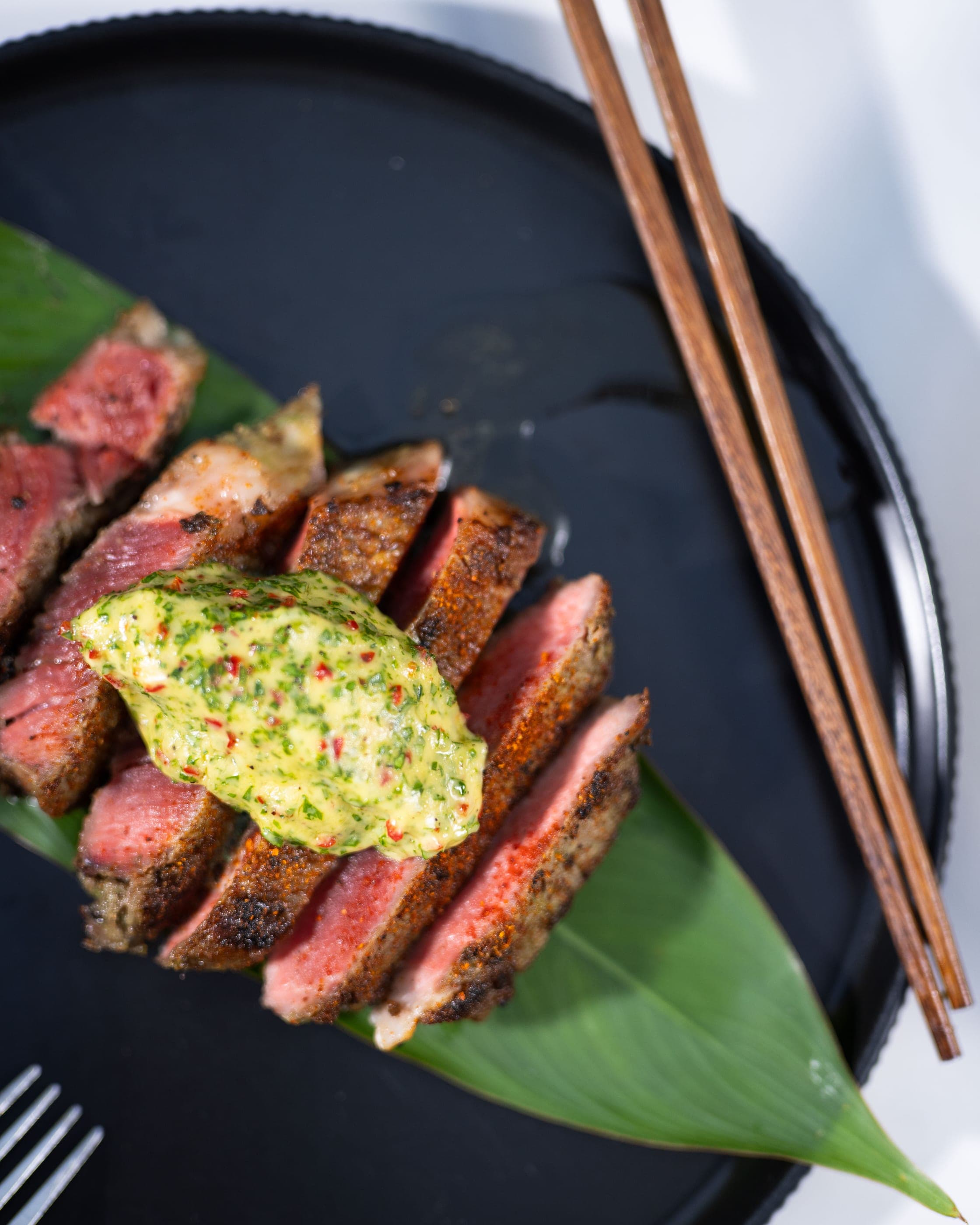 Sliced seared steak under a green herb butter on a dark plate