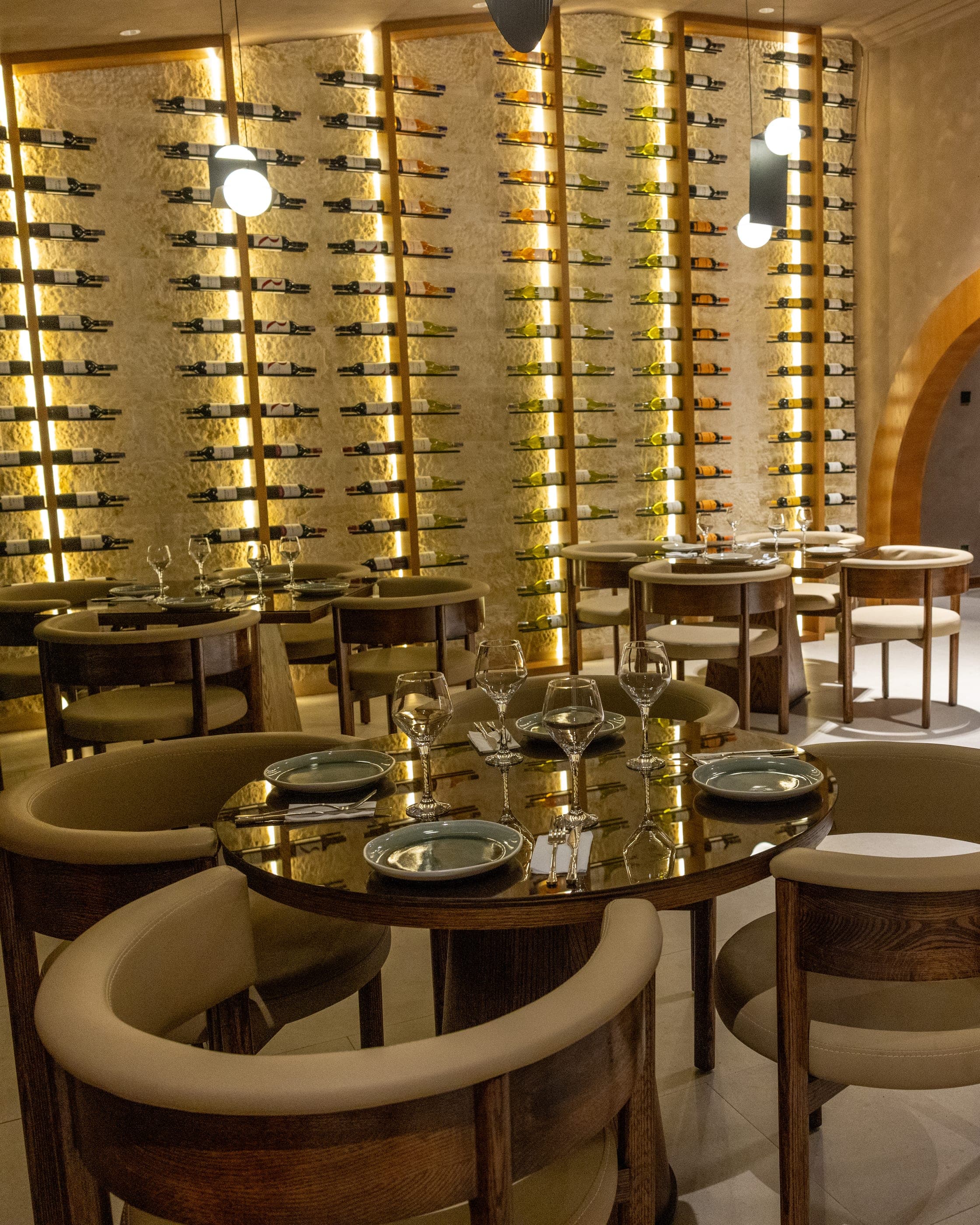 The dining room from the far end, tables laid under pendants beside the lit wine wall